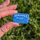 A hand holding a small blue rectangular tin with embossed white text reading 'SARDINES IN OLIVE OIL' against a blurred green outdoor background.
